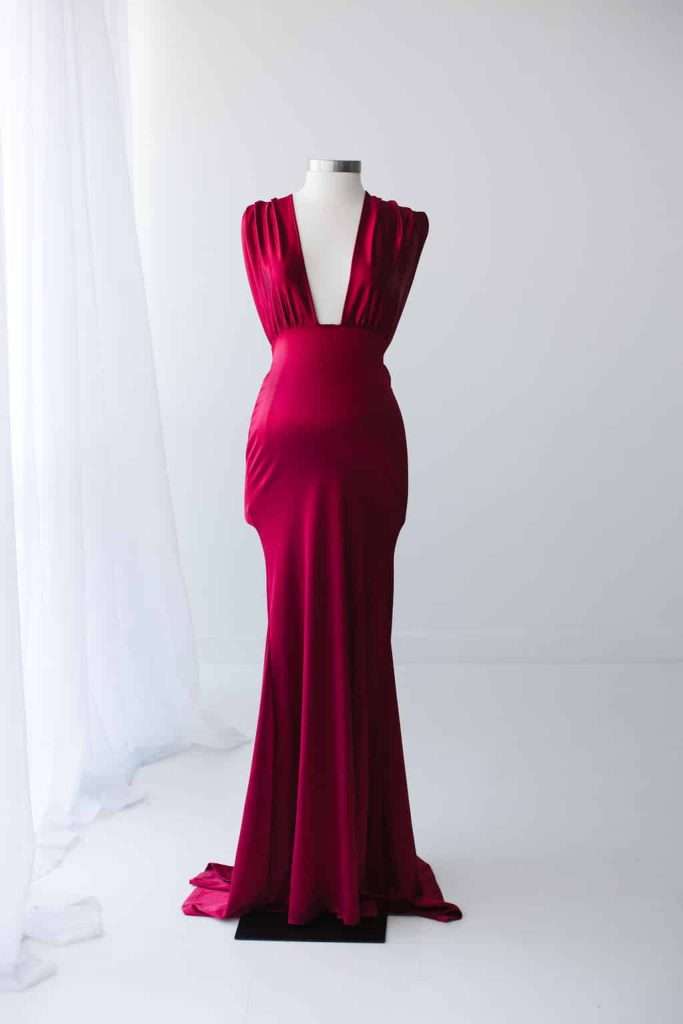 A deep red, sleeveless evening gown with a plunging neckline is displayed on a white mannequin in a minimalist, white room.