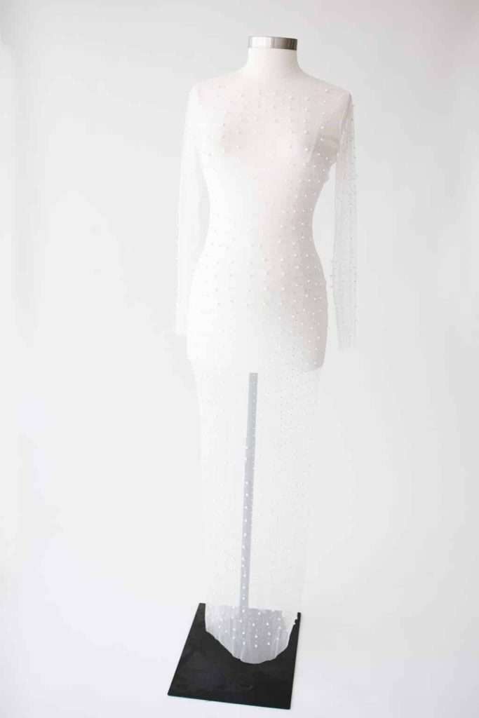 A sheer white dress with long sleeves and pearl embellishments displayed on a dress form against a plain white background.