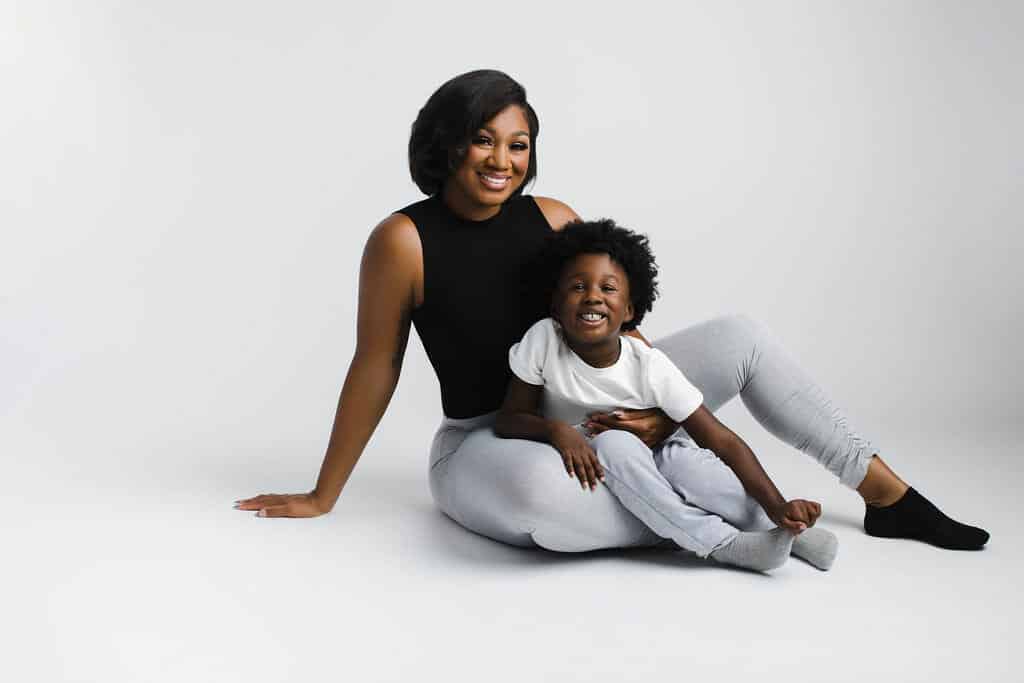 A woman and a young child sit on the floor together against a plain white background, both smiling at the camera.