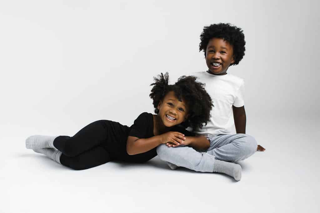 Two children sit on the floor against a plain white background, smiling. One child is lying on their side, while the other sits cross-legged beside them.