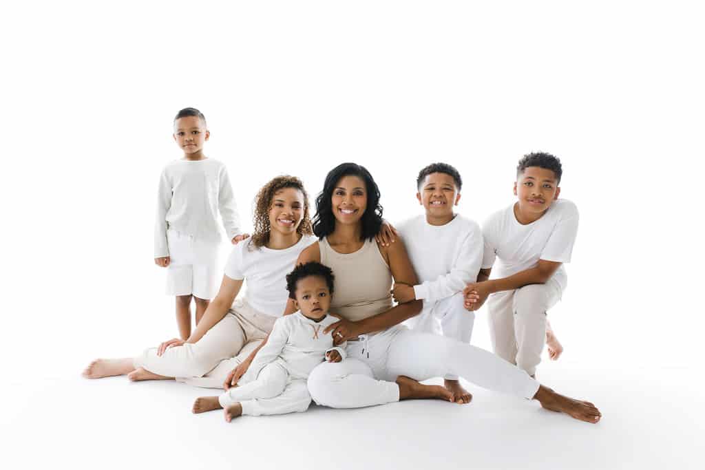 A woman sits on the floor with five children, all wearing white or light-colored clothing, against a plain white background.
