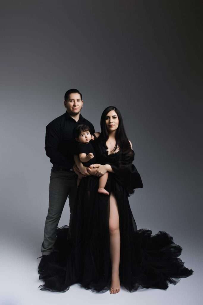 A man stands beside a seated woman in a flowing black dress holding a toddler. All three face forward against a plain, neutral background.
