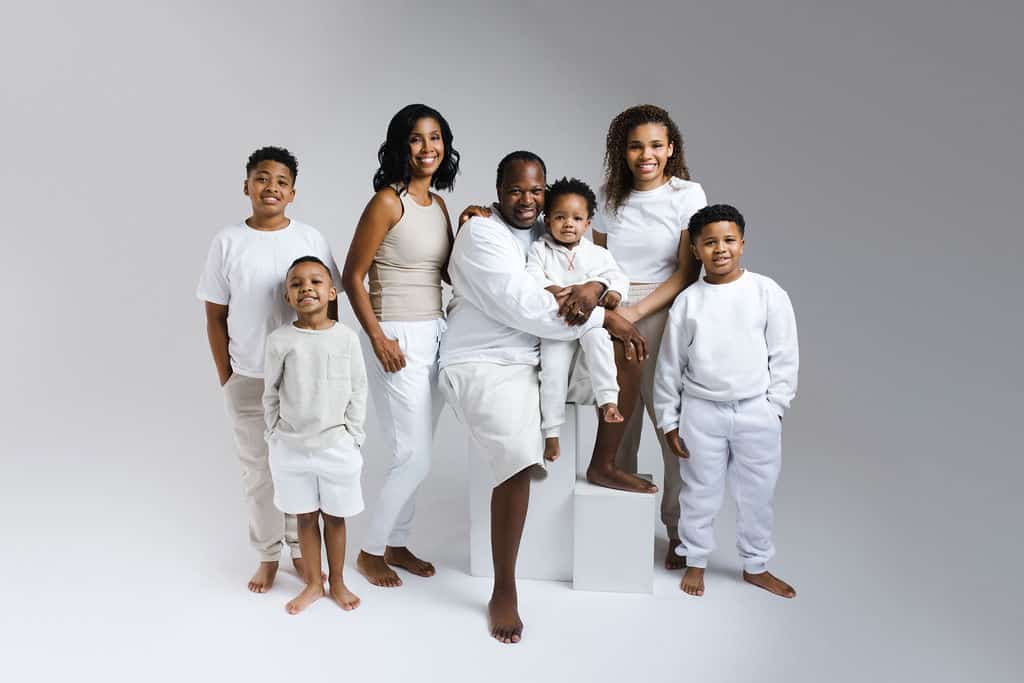 A family of seven, all barefoot and dressed in white and beige, poses together against a plain light background.