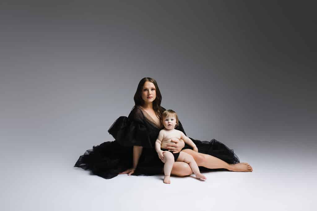 Woman in a black dress sits on the floor with a young child on her lap, both looking at the camera, against a plain gray background.