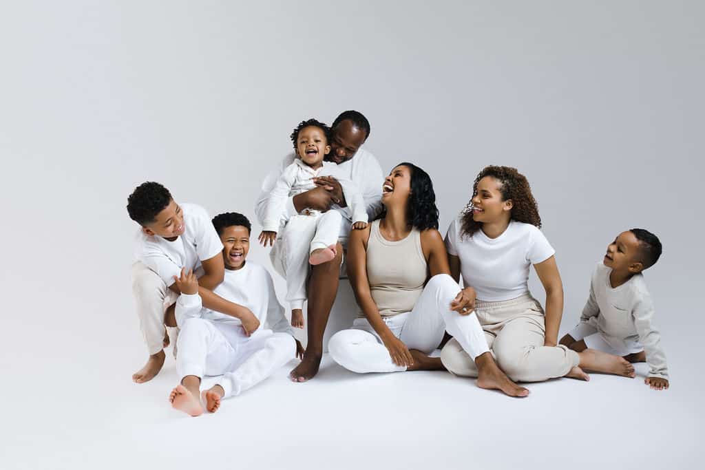 Six people, including children and adults, sit and laugh together on the floor against a plain white background, all wearing white or neutral clothing.