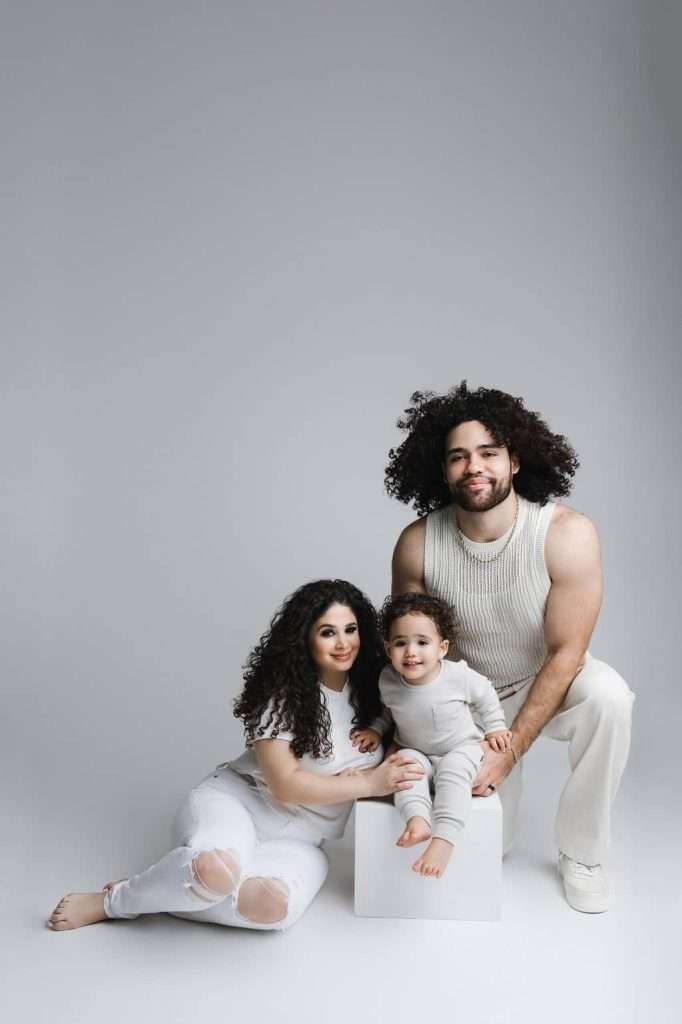 A woman, a man, and a young child with curly hair pose together against a plain light gray background, all dressed in white clothing.
