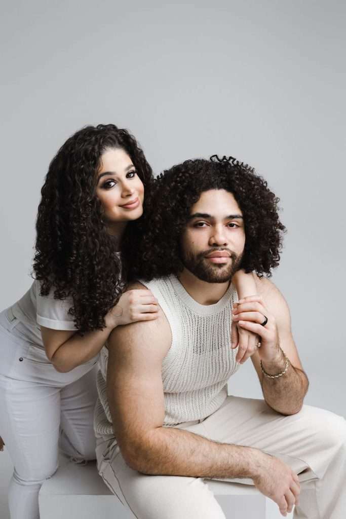 A woman with curly hair stands behind a seated man with curly hair, both wearing light-colored, casual outfits, against a plain gray background.