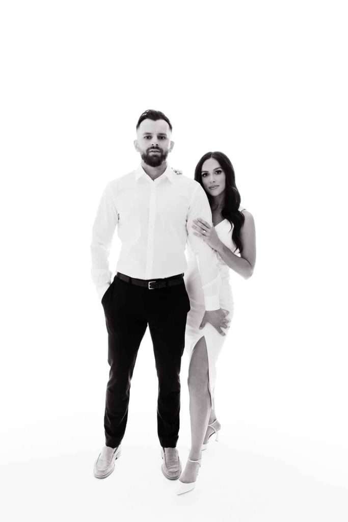 A man in a white shirt and dark pants stands with his hands in his pockets. A woman in a white dress with a slit stands beside him, her hand resting on his shoulder.