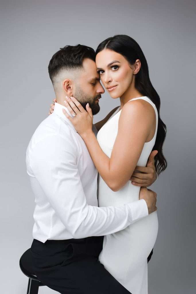 A man in a white shirt and dark pants sits on a stool while a woman in a sleeveless white dress sits on his lap, both looking at the camera with neutral expressions.