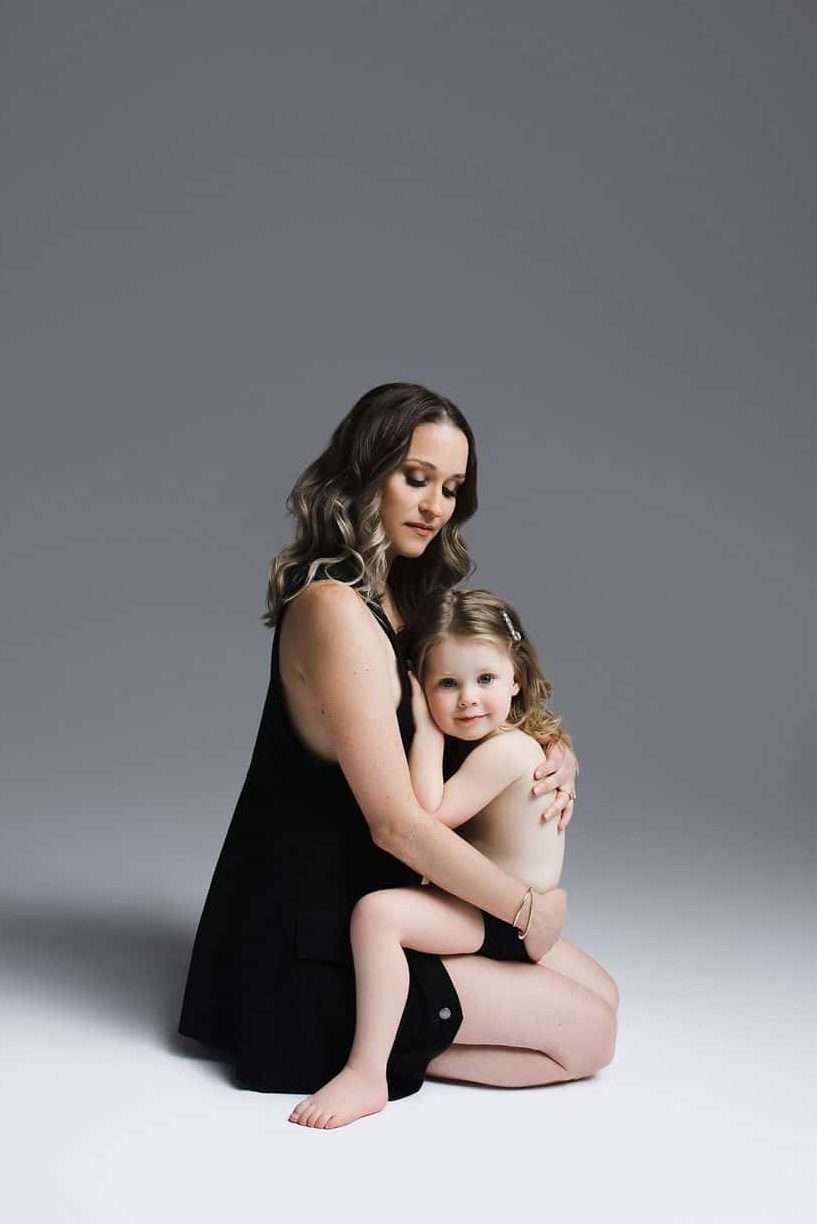 A woman sits on the floor holding a young child in her lap. Both are dressed in black and pose against a plain gray background.