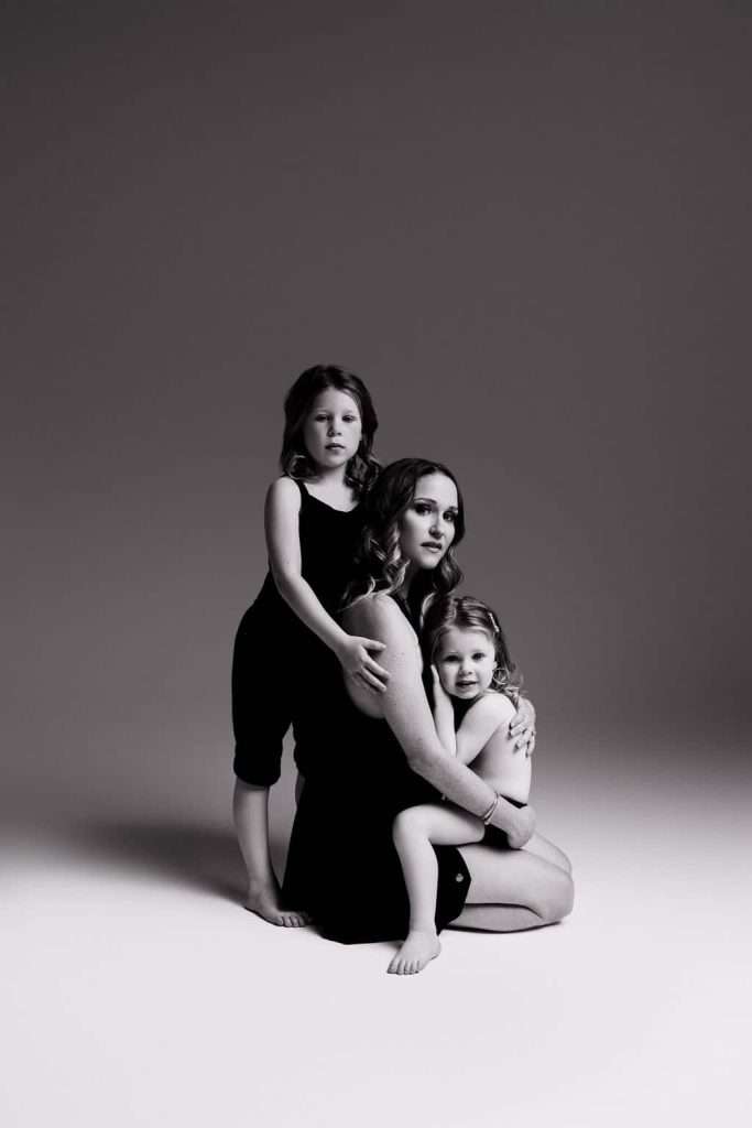 A woman sits on the floor embracing two young children, all wearing dark clothing, against a plain studio backdrop in black and white.