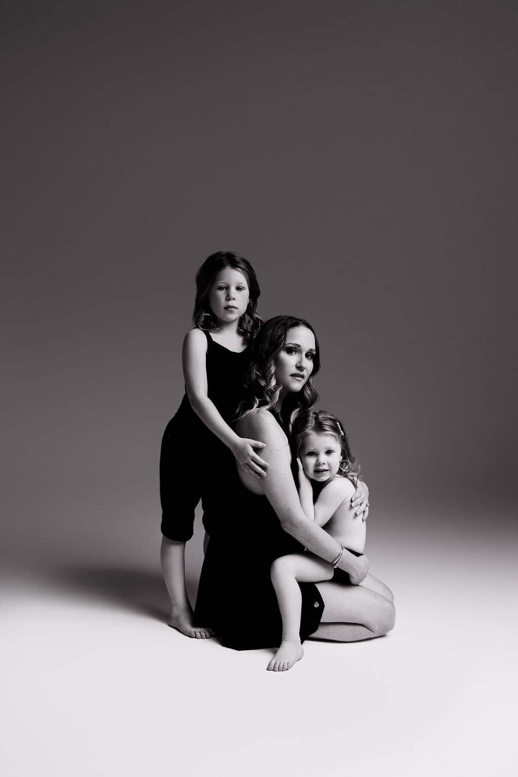 A woman sits on the floor embracing two young children, all wearing dark clothing, against a plain studio backdrop in black and white.