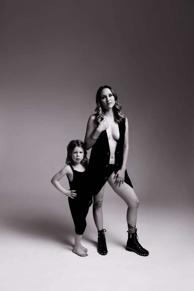 A woman and a young girl stand together in a studio. The woman wears an open vest and boots; the girl wears a sleeveless top and leggings. Both face the camera with neutral expressions.