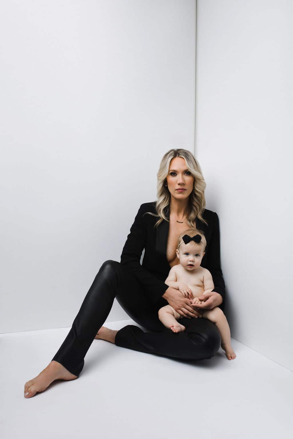 A woman in a black outfit sits on the floor against a white wall, holding a baby wearing only a black bow headband.