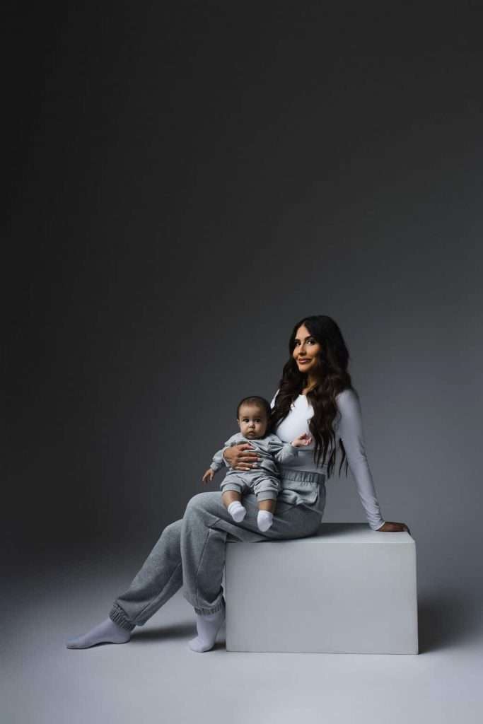 Woman in gray sweatpants and a white top sits on a white block holding a baby in striped clothing; both face forward against a plain gray background with soft lighting.