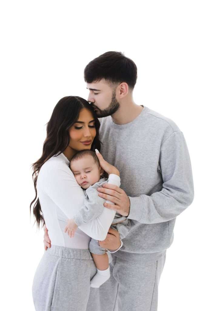 A man kisses a woman’s forehead as she holds a sleeping baby; all three wear light gray clothing against a white background.