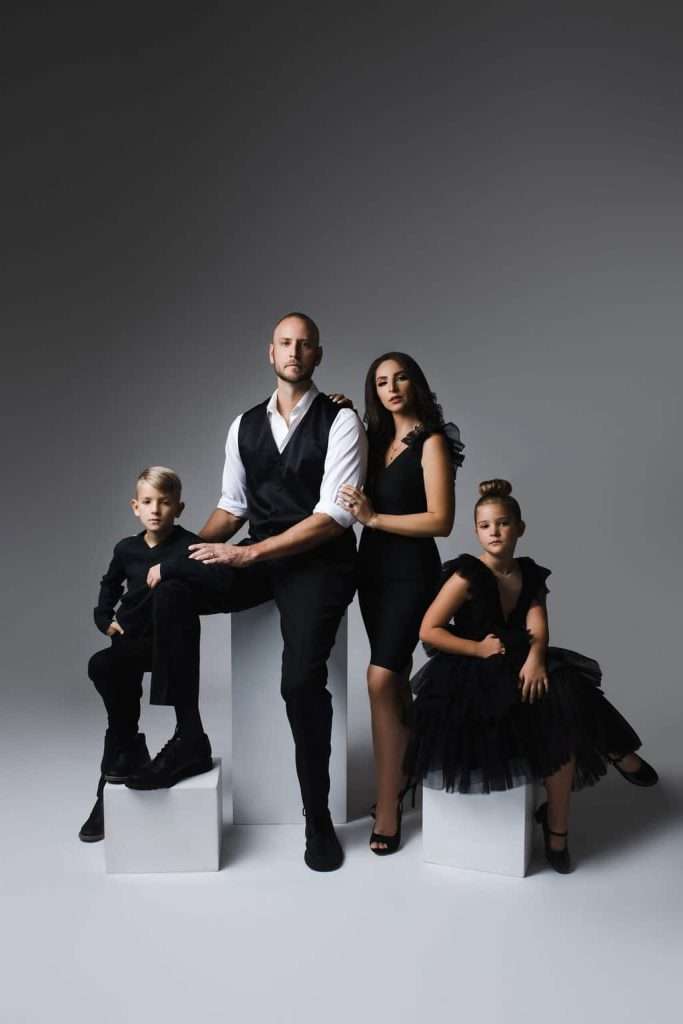 Four people in black formal attire pose together against a plain gray backdrop; two adults stand in the center, with one boy seated and one girl in a black dress sitting on either side.