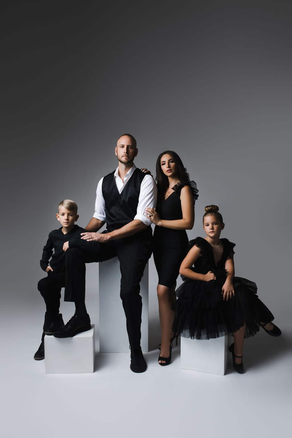 Four people in black formal attire pose together against a plain gray backdrop; two adults stand in the center, with one boy seated and one girl in a black dress sitting on either side.