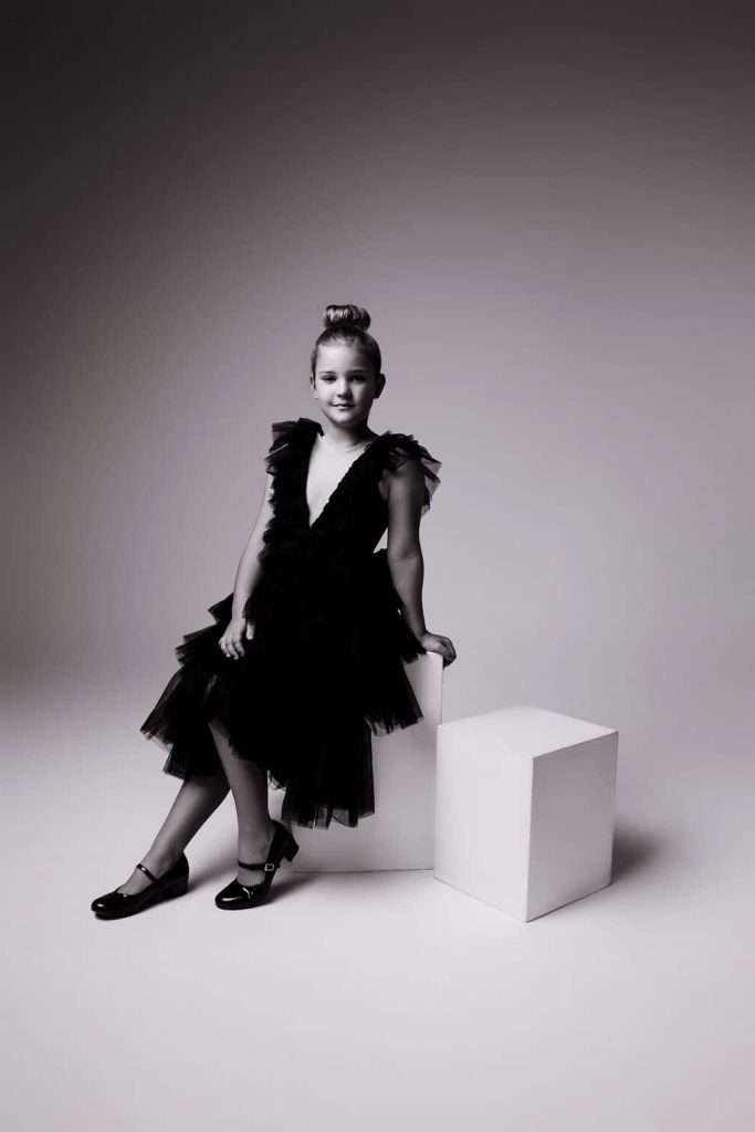 Young girl in a black dress with tulle skirt sits on a white cube in a studio with a plain background.