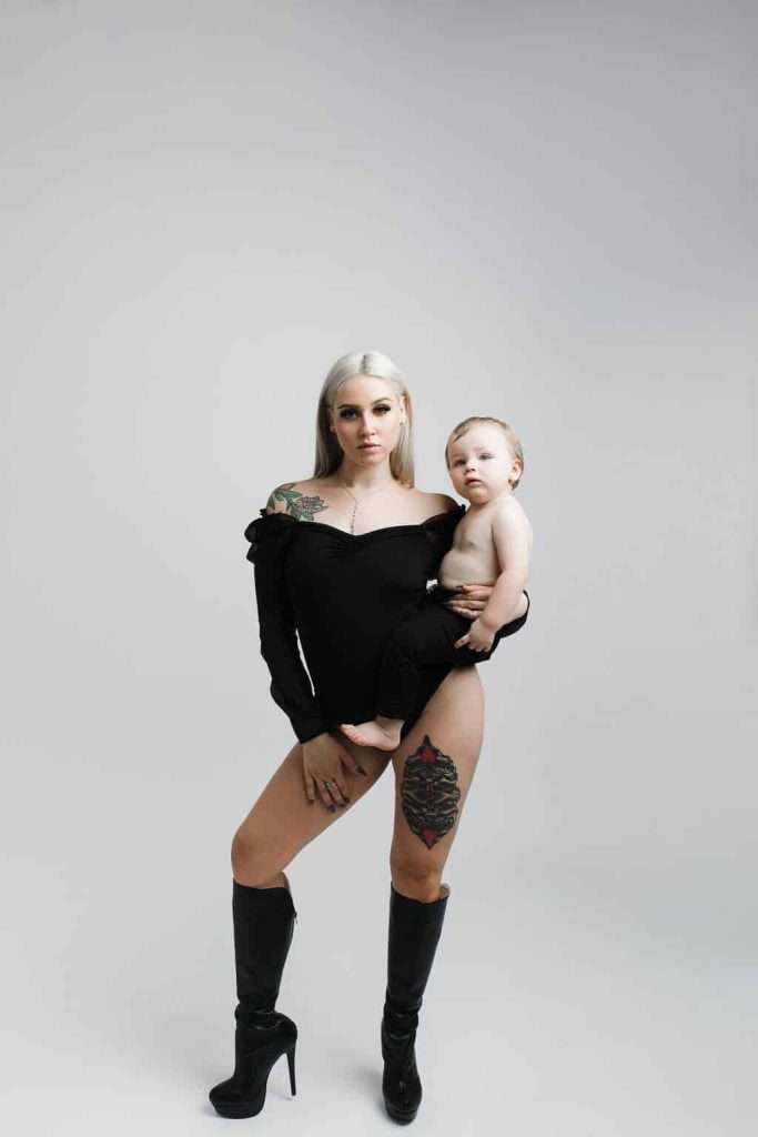 Woman in a black outfit and high-heeled boots stands holding a baby. Both have tattoos visible. White background.