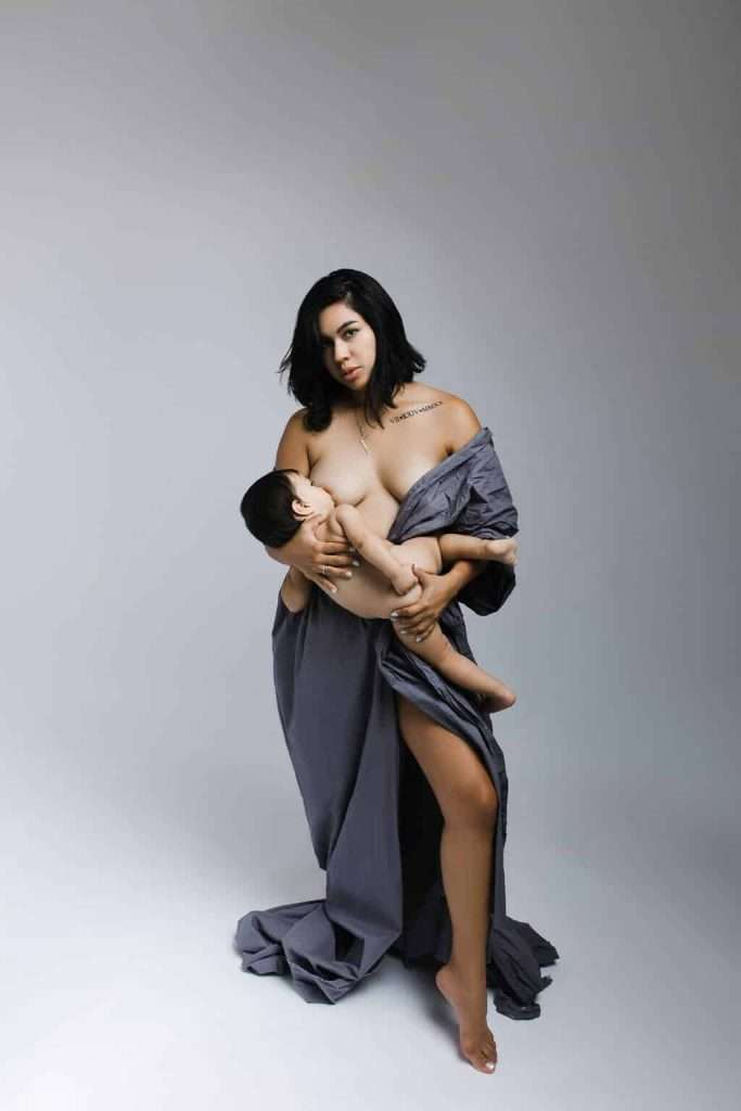 Woman wrapped in a gray sheet standing barefoot, breastfeeding a baby while looking at the camera against a plain background.