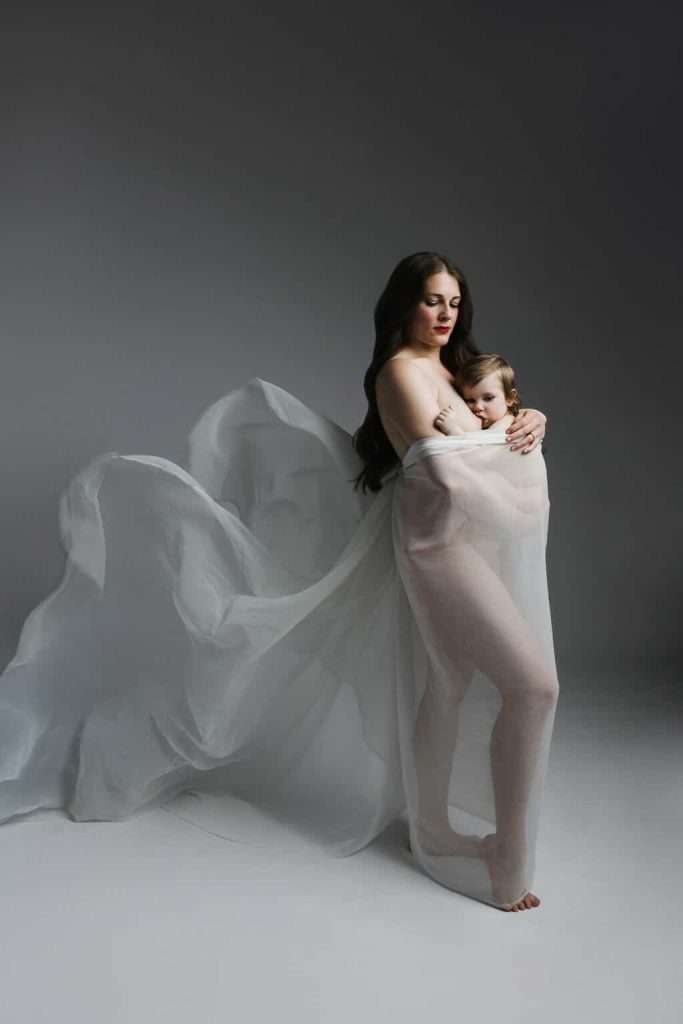 A woman and a child are standing together wrapped in sheer, flowing white fabric against a plain background.