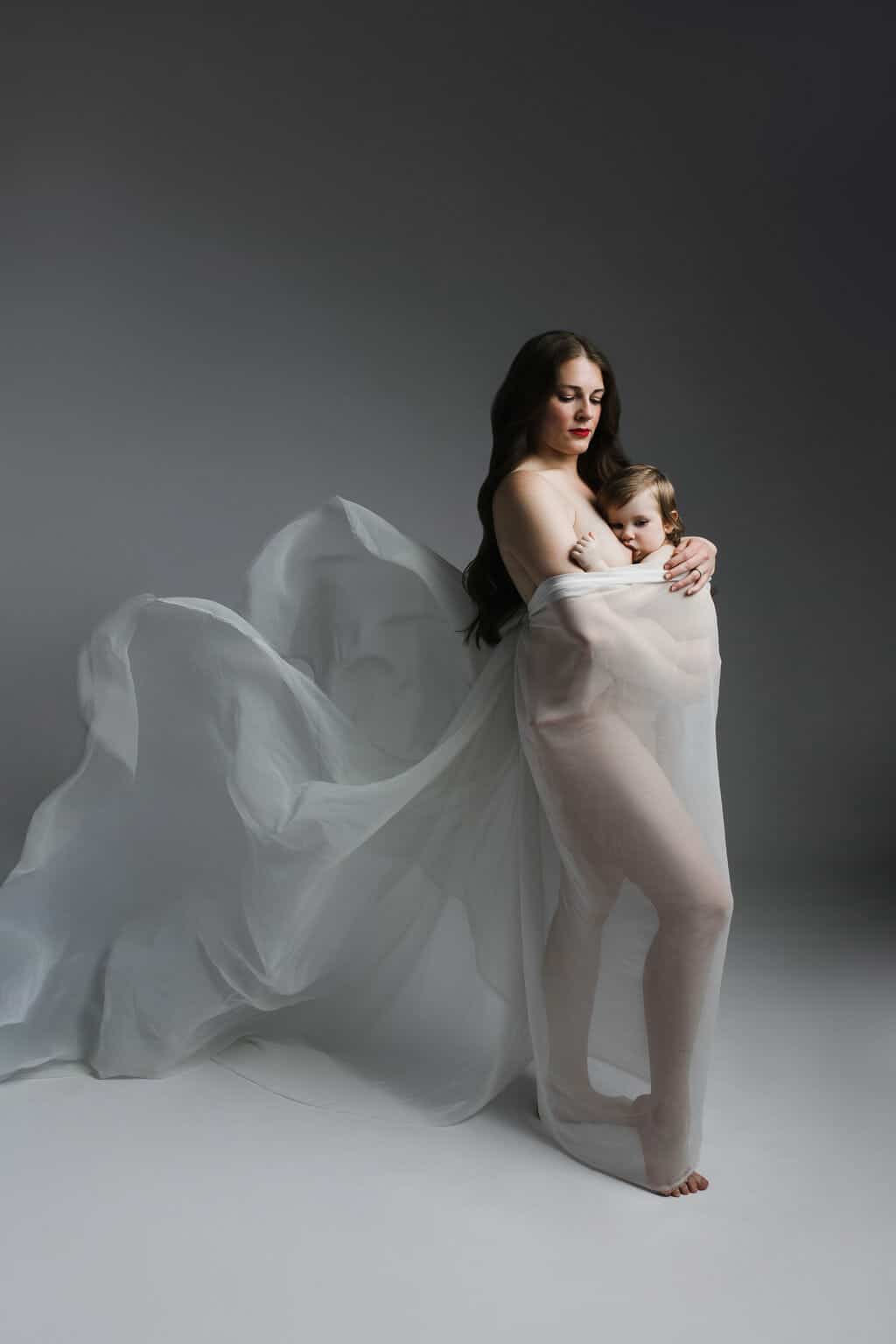 A woman and a child are standing together wrapped in sheer, flowing white fabric against a plain background.