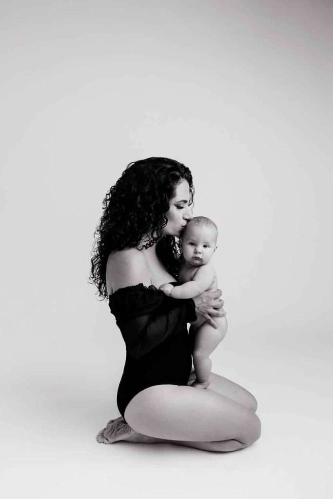 Woman in a black off-shoulder outfit kneels on the floor, holding and kissing a baby. The photo is in black and white.