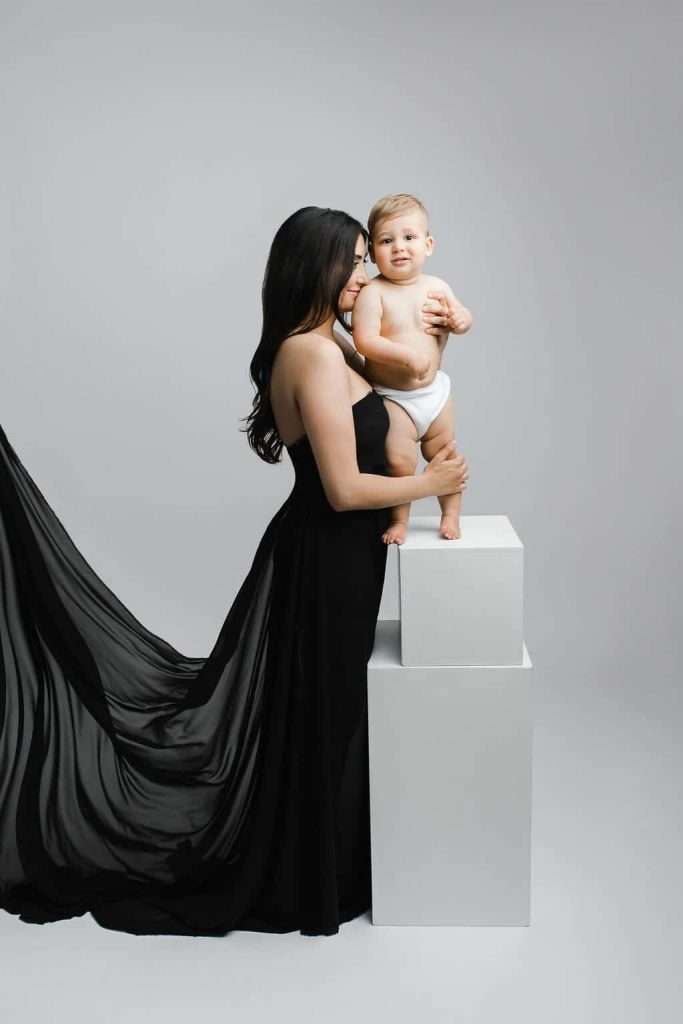Woman in a black dress holds a baby wearing a diaper, both standing next to white blocks against a gray backdrop.