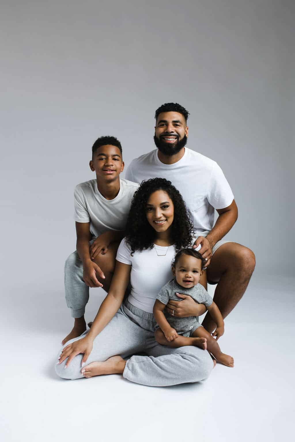 A family of four poses against a plain backdrop, with two adults and two children dressed in casual clothing, all smiling at the camera.