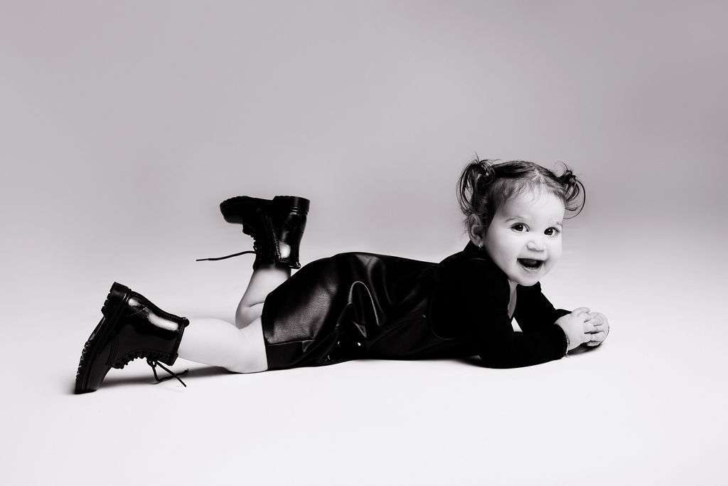 Toddler with pigtails lies on stomach, smiling, wearing a black outfit and lace-up boots against a plain background.