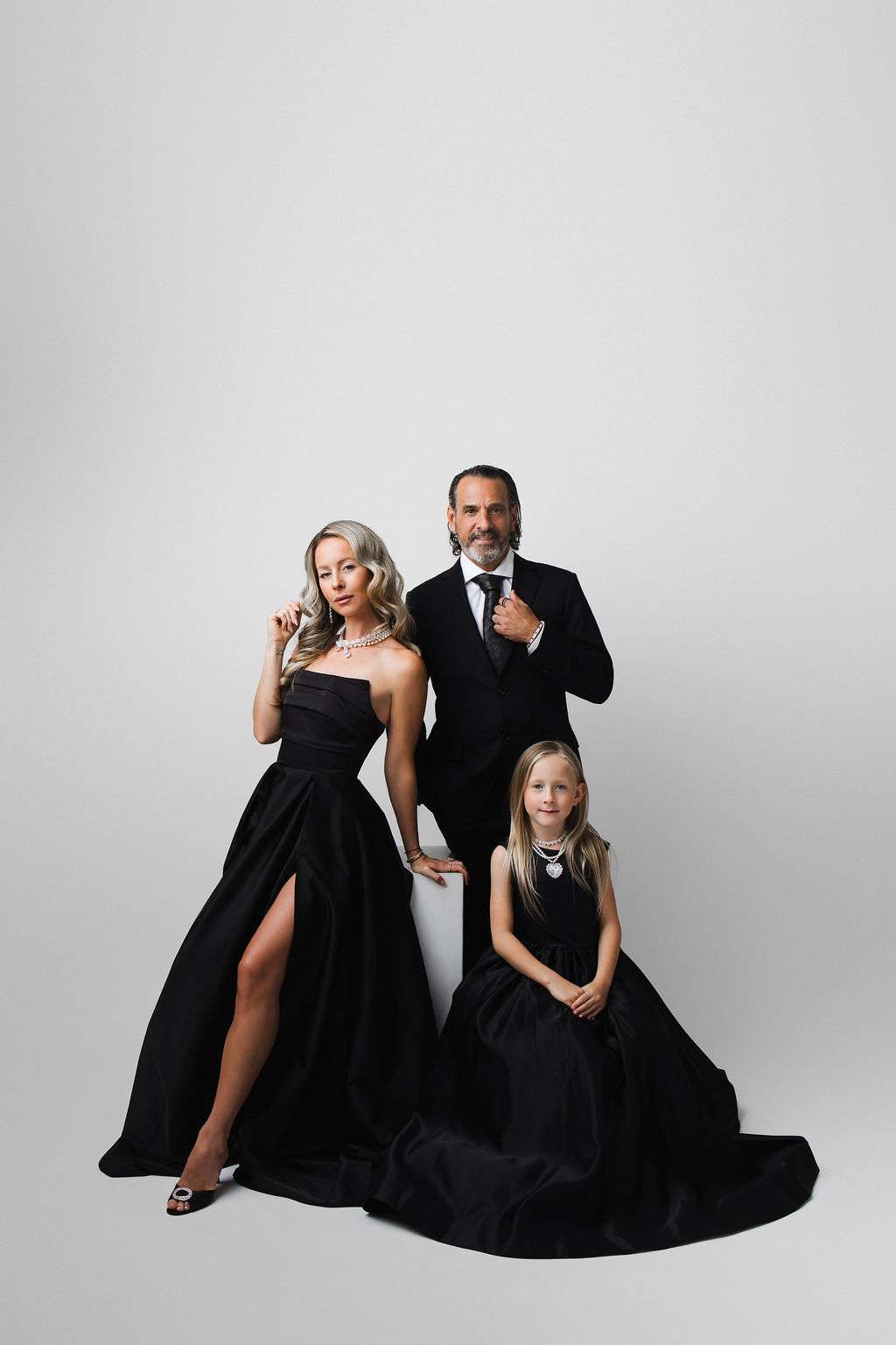 Three people pose against a plain white background; two adults in formal black attire stand, while a child in a black dress sits on the floor, all looking at the camera.
