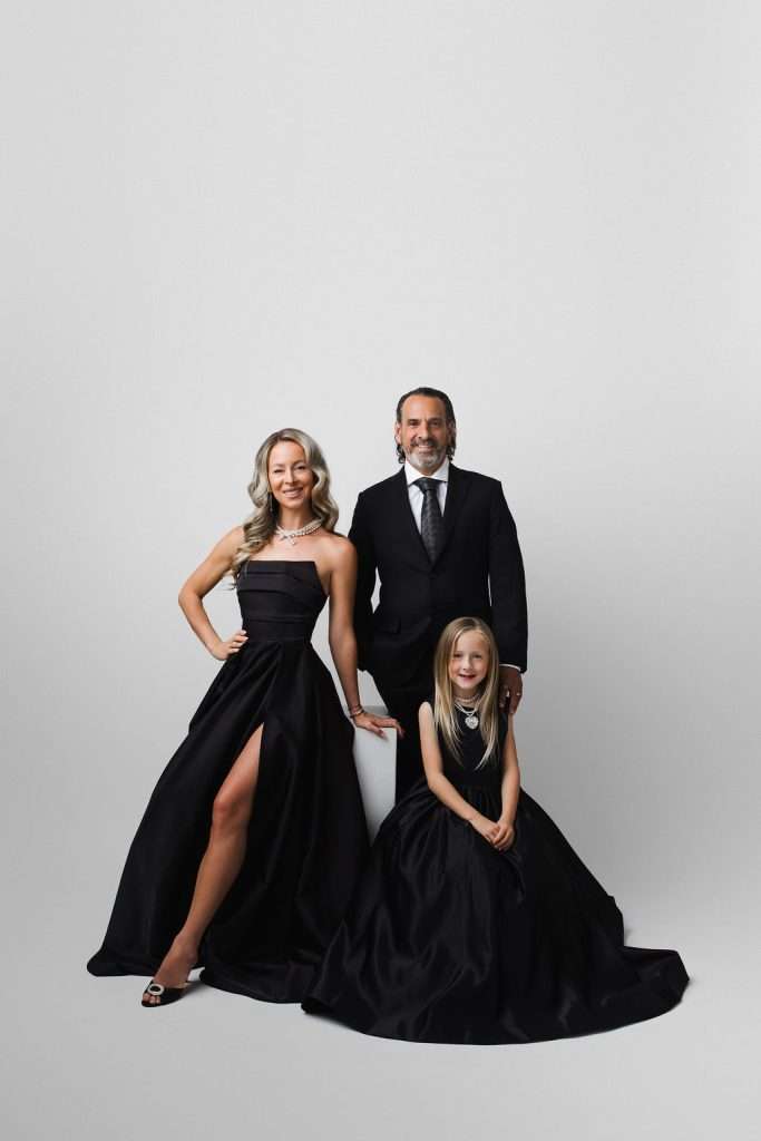 Three people pose in formal black attire against a plain white background; an adult woman stands, an adult man stands behind, and a young girl sits in front.
