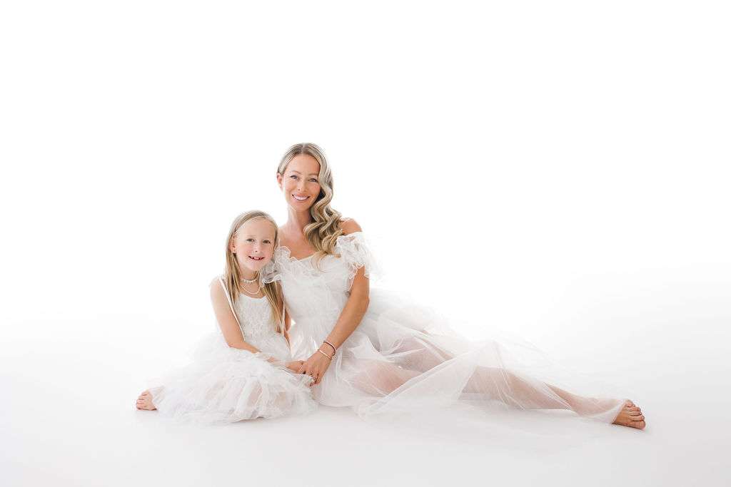 A woman and a young girl, both in white dresses, sit on the floor against a white background, smiling at the camera.