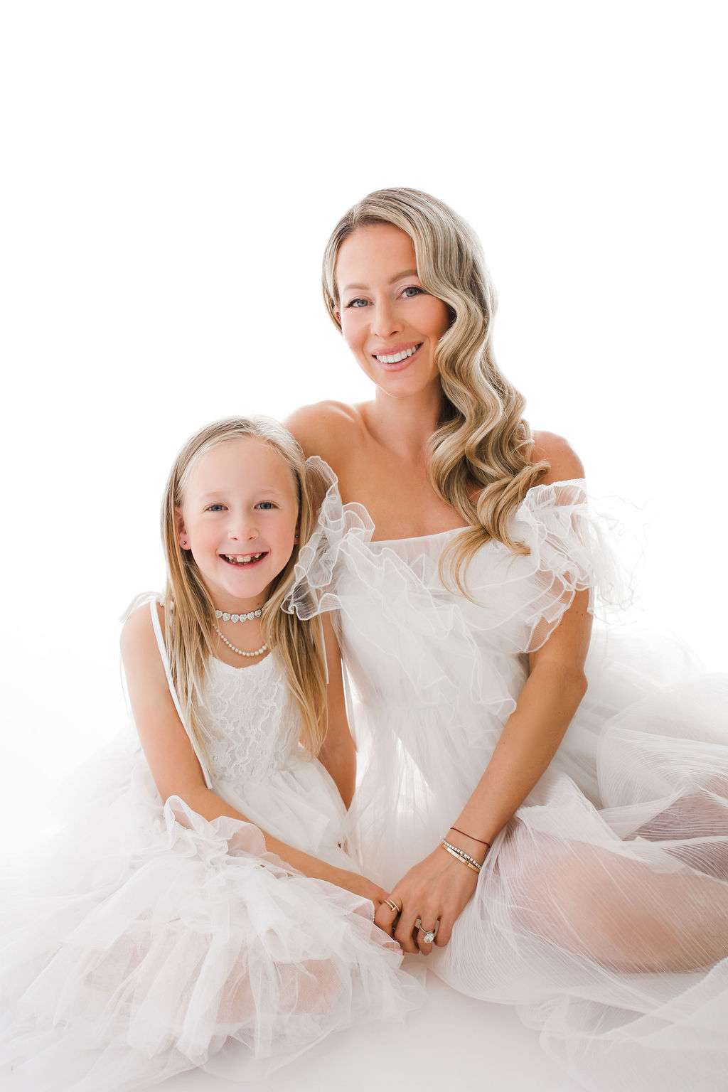 A woman and a young girl, both wearing white tulle dresses, sit together and smile against a plain white background.