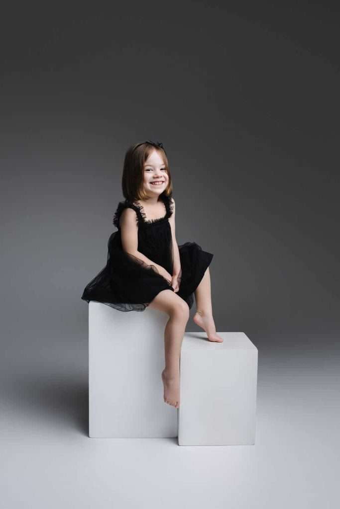 Young girl in a black dress sits barefoot on white cubes against a plain gray background, smiling at the camera.
