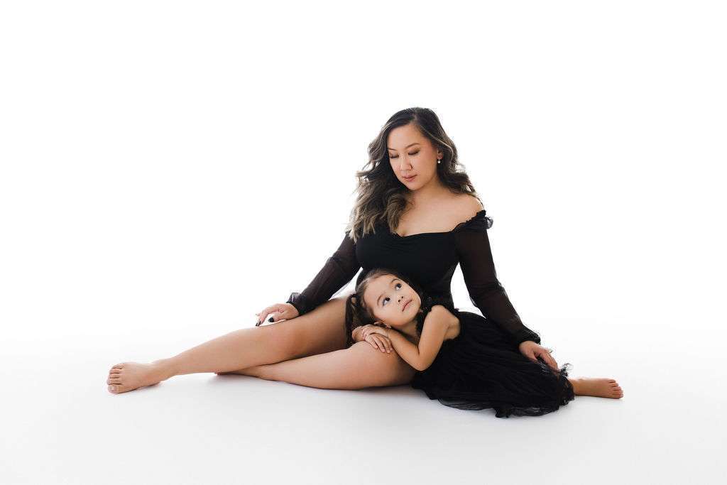 A woman in a black dress sits on the floor with a young girl in a matching dress resting her head on the woman’s leg against a white background.