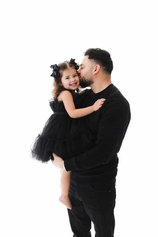 Man in black holds a smiling young girl in a black dress while kissing her forehead, both standing against a white background.