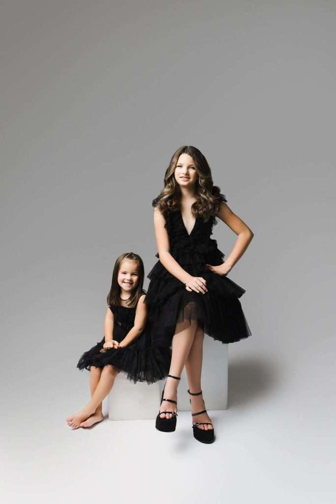 Two young girls in black dresses pose together in a studio with a light gray background. One sits on a white cube while the other sits on the floor, both smiling at the camera.