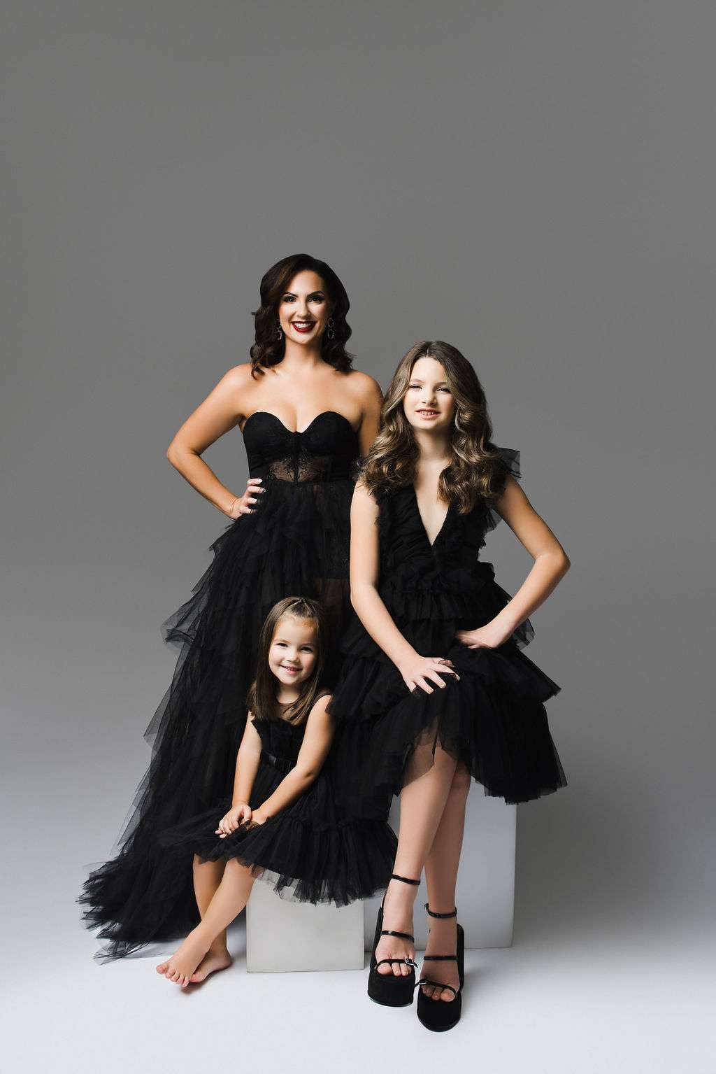 Three females wearing black dresses pose together in a studio with a plain gray background. Two are standing or seated on a block, and one sits on the floor in front.