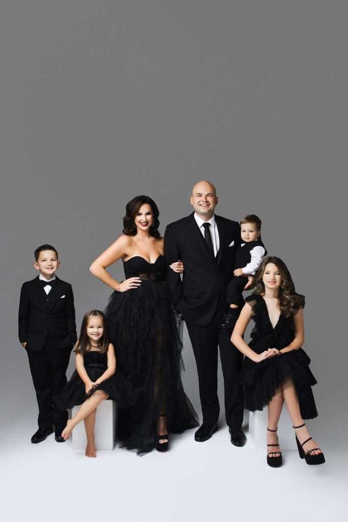 A family of six in formal black attire poses together against a plain gray background. The adults and children are dressed in coordinated outfits, some seated and some standing.