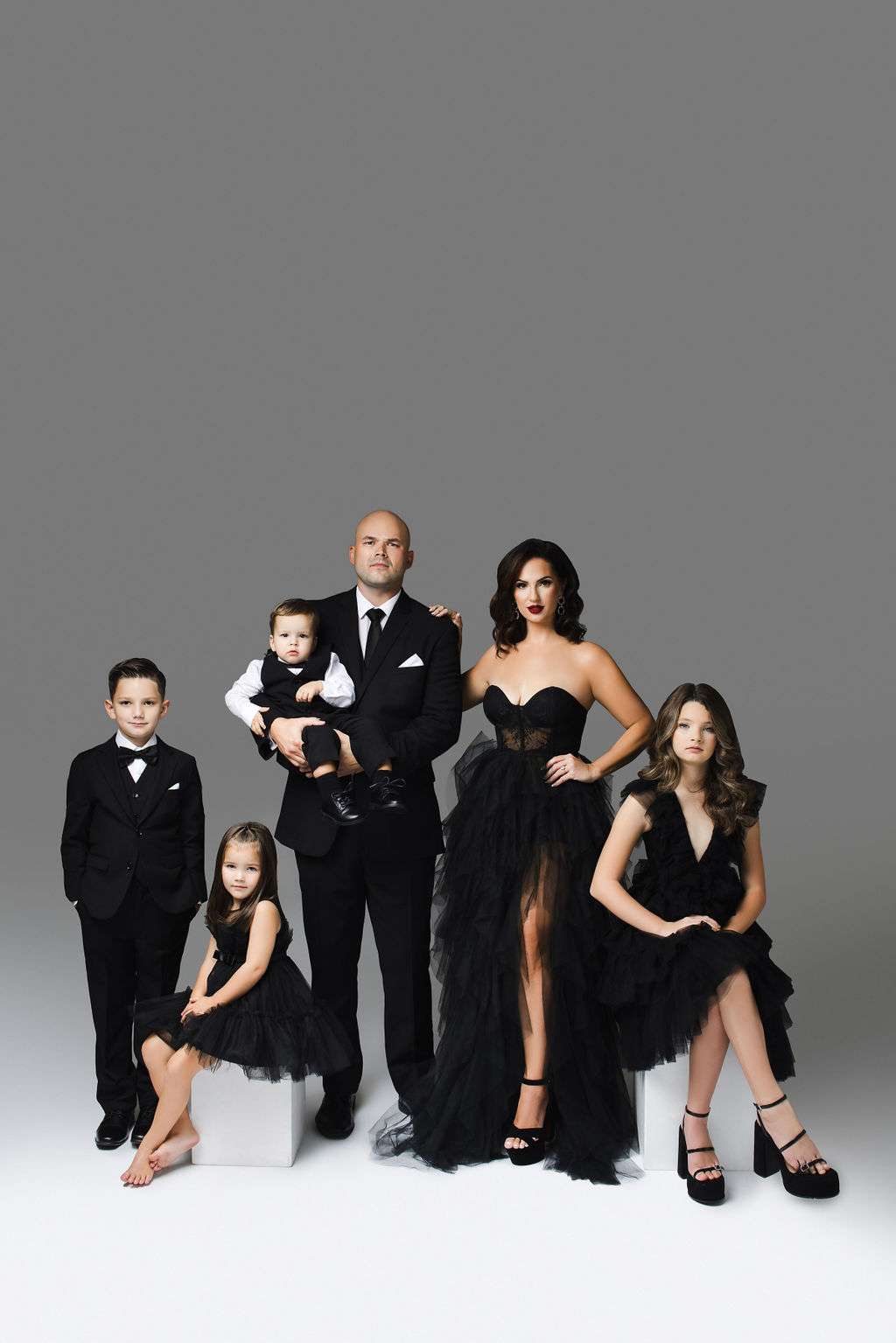 A family of six poses in formal black attire against a gray gradient background; the adults stand while the children are seated or held.