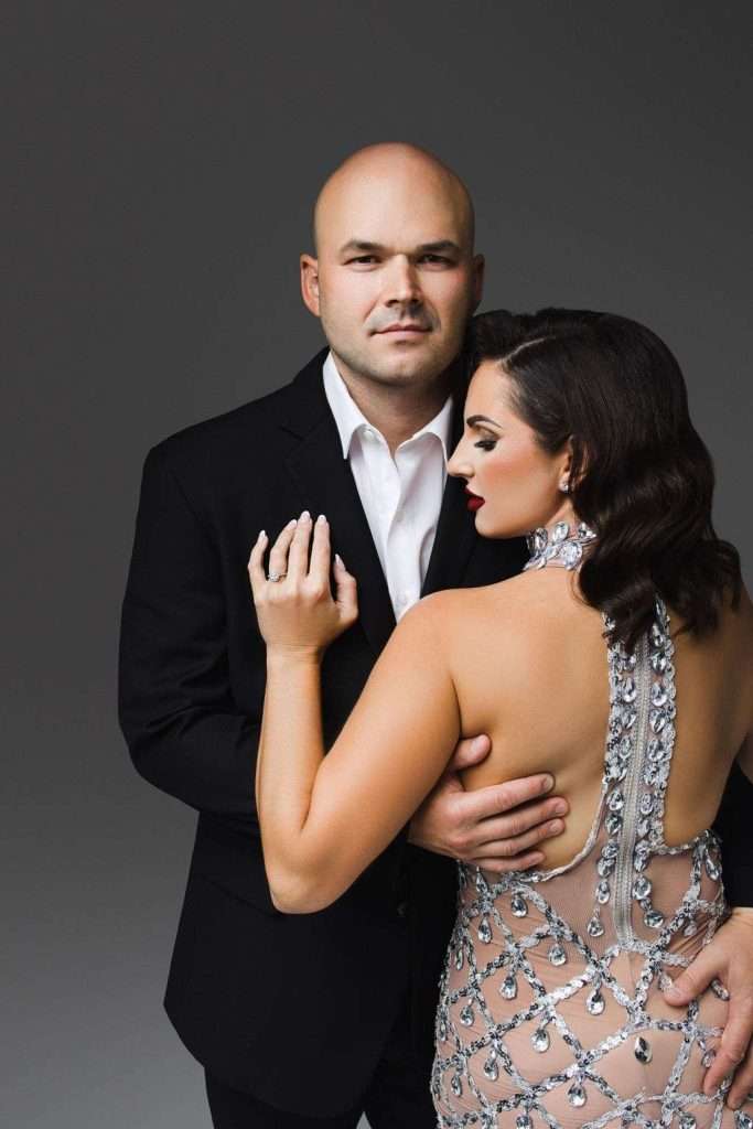 A man in a black suit stands next to a woman in a jeweled, backless dress. She faces him, and he holds her waist while looking at the camera.