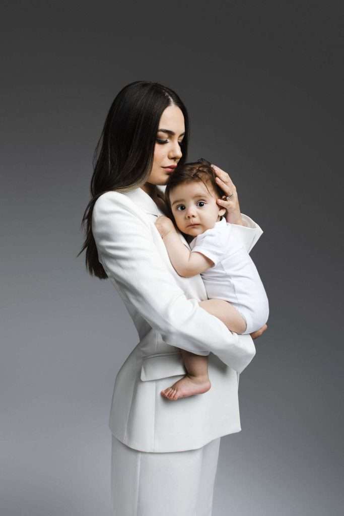 A woman in a white suit holds a baby dressed in white against her chest. She gently supports the baby’s head while the baby looks forward. The background is plain and gray.