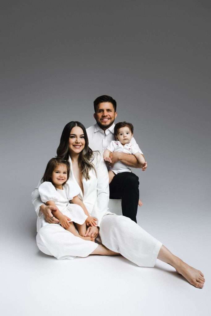A family of four poses in a studio wearing white clothes, with two adults and two young children sitting together against a plain gray gradient background.