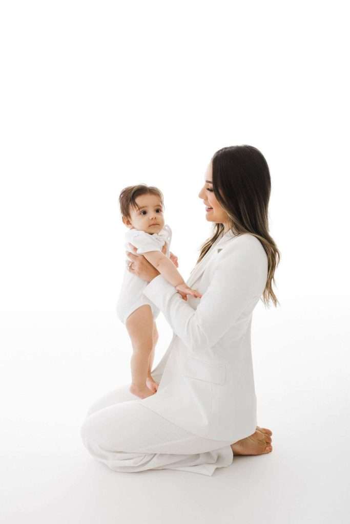 Woman in a white suit kneeling on the ground, holding a baby dressed in a white onesie, both facing each other against a white background.