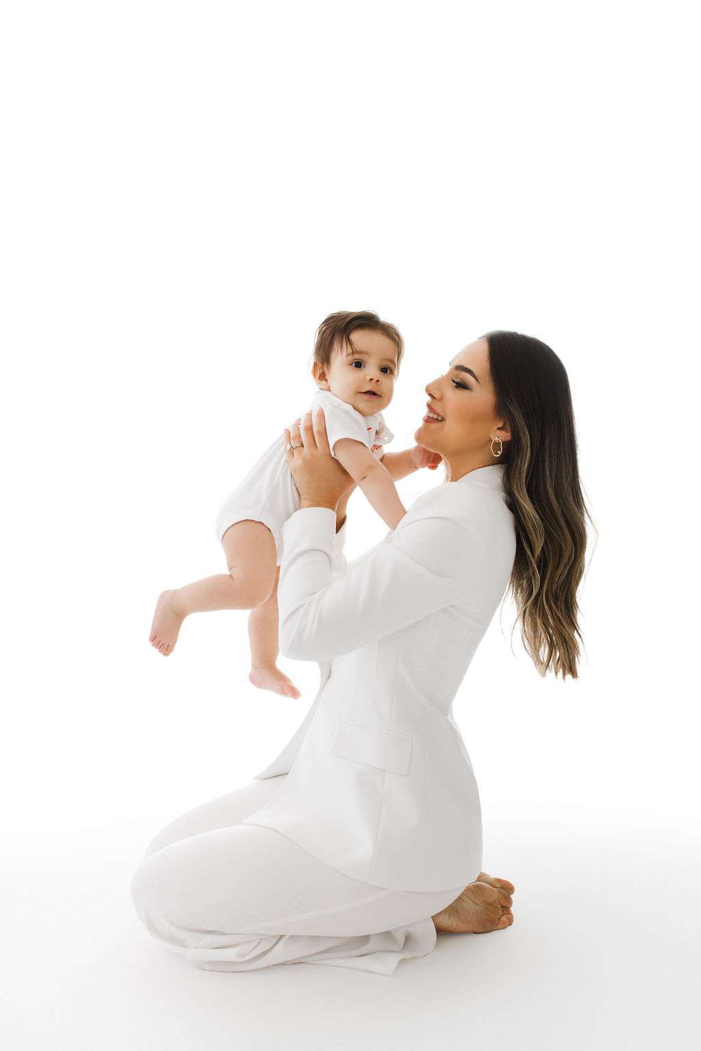 Woman in white kneels on the floor, holding a baby up in front of her. Both are looking forward against a plain white background.