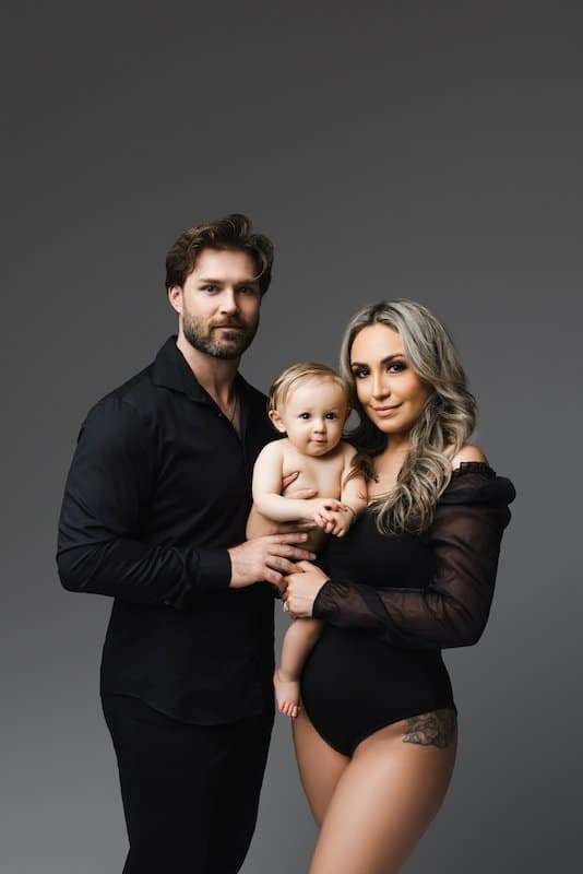 A man and woman dressed in black pose together with a baby in front of a neutral gray background.