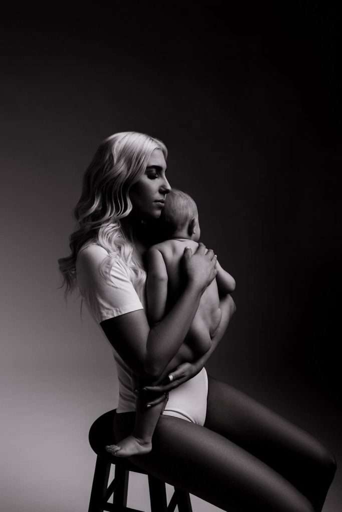 A woman sits on a stool holding a small baby against her chest. The photo is in black and white with dramatic lighting and shadow.