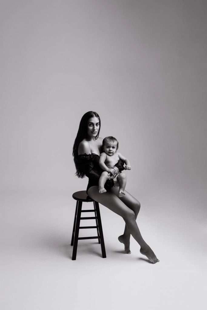 A woman in a black outfit sits on a stool holding a smiling baby, both facing the camera in a minimalist studio setting.